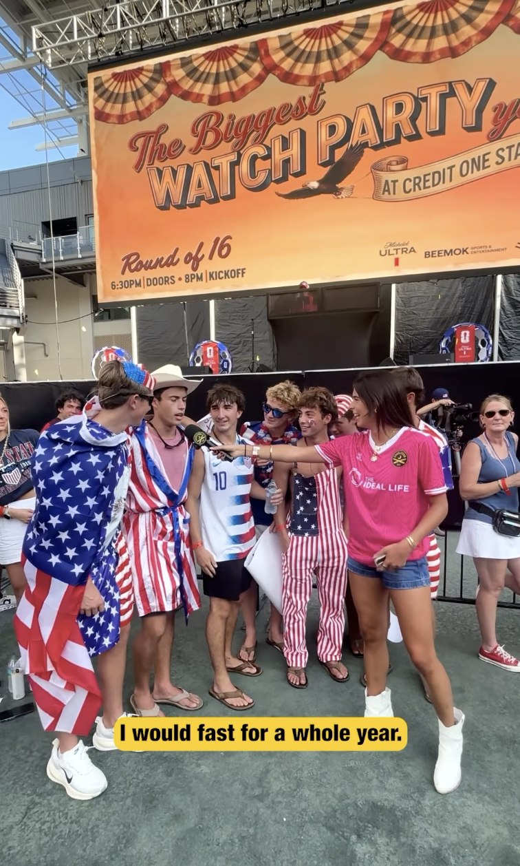 A group of young people dressed in patriotic American outfits being interviewed at a festive event, with a large banner in the background advertising 'The Biggest Watch Party' event.
