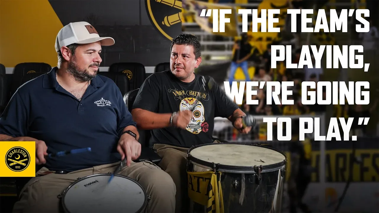 Two men sitting in chairs with drums, one holding drumsticks, at a sports bar or game room. One man wearing a navy polo shirt and cap, the other wearing a black t-shirt with a skull and rose design. Large white text overlay says, 'If the team’s playing, we’re going to play.' A Charleston logo is in the bottom left corner.