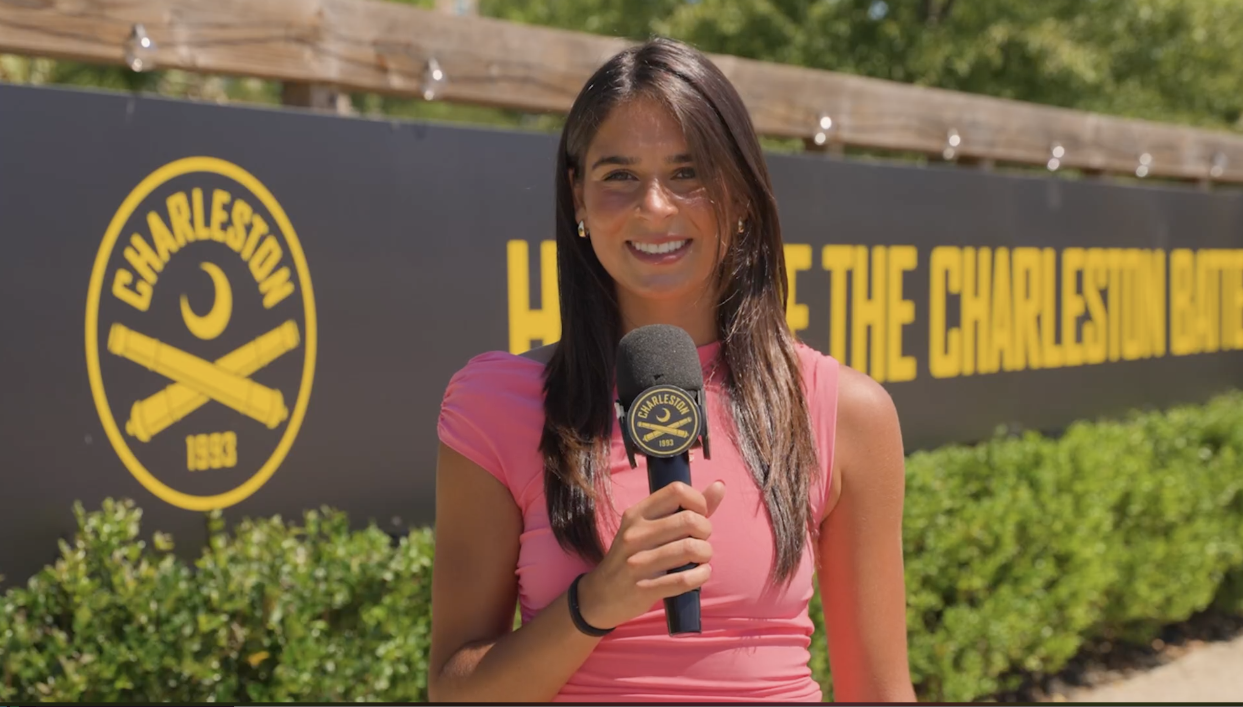 A woman with long dark hair holding a microphone with the Charleston logo, standing outdoors near a large Charleston sign with yellow text and a black background, surrounded by green foliage.