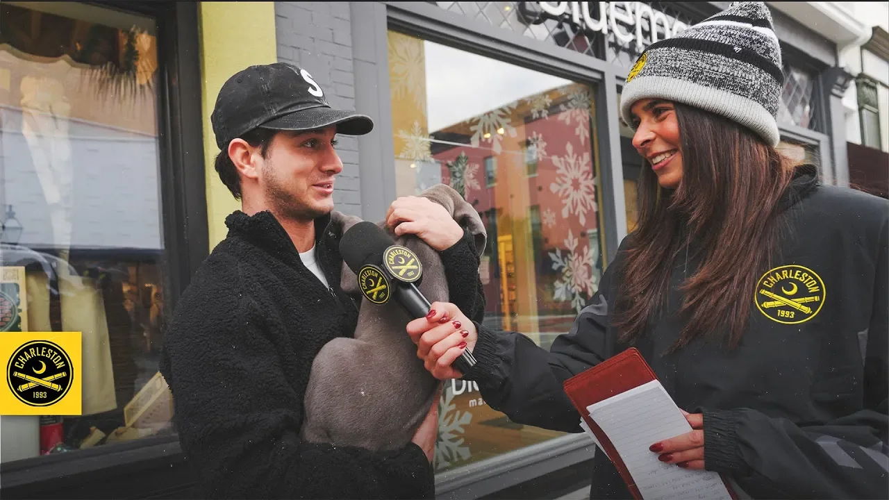 A woman wearing a Charleston jacket and a striped beanie holds a microphone while interviewing a man holding a stuffed animal outside a storefront decorated with winter holiday decor.