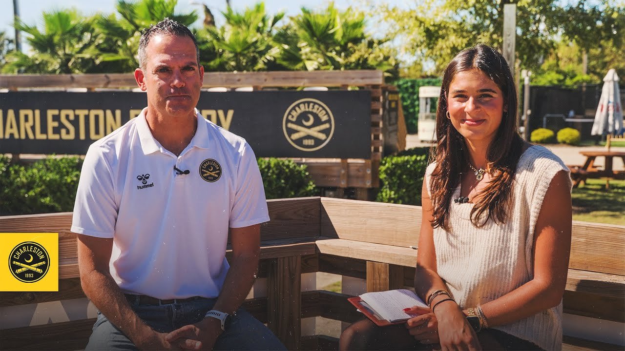 A man and a woman sitting outdoors on a wooden bench with greenery and a Charleston Battery sign in the background. The man is wearing a white polo shirt and the woman has long brown hair, wearing a light-colored sleeveless top, holding a notebook.