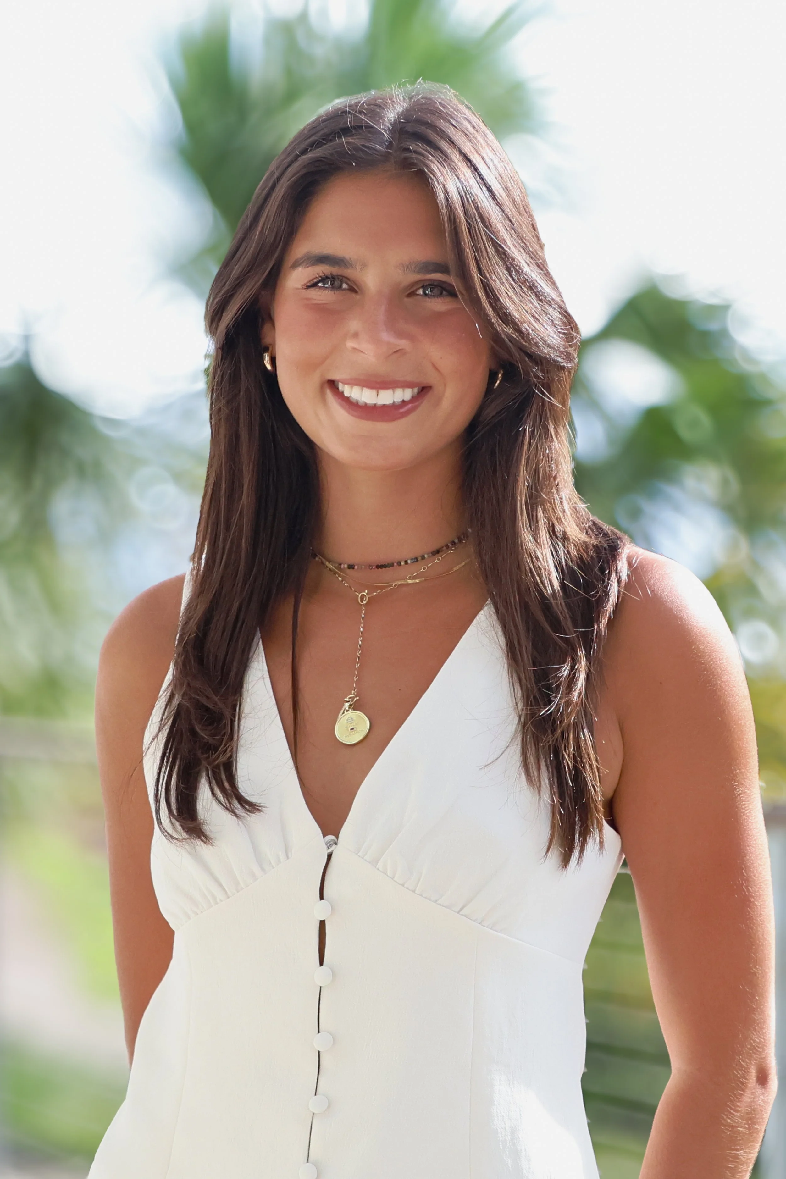 A young woman with long brown hair, smiling outdoors with blurred green foliage in the background, wearing a white sleeveless dress with buttons and layered necklaces.
