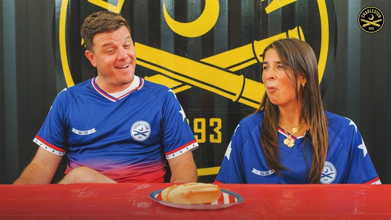 A man and woman sitting at a table, wearing matching blue Charleston soccer jerseys with red, white, and blue accents, in front of a black and yellow Charleston Soccer logo. They are smiling and looking at each other, with a hot dog on a plate on the table.