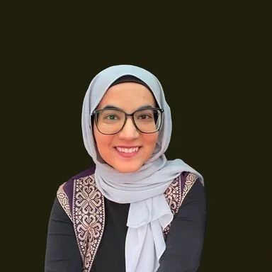 A woman wearing glasses and a light gray hijab, smiling at the camera with a dark background.