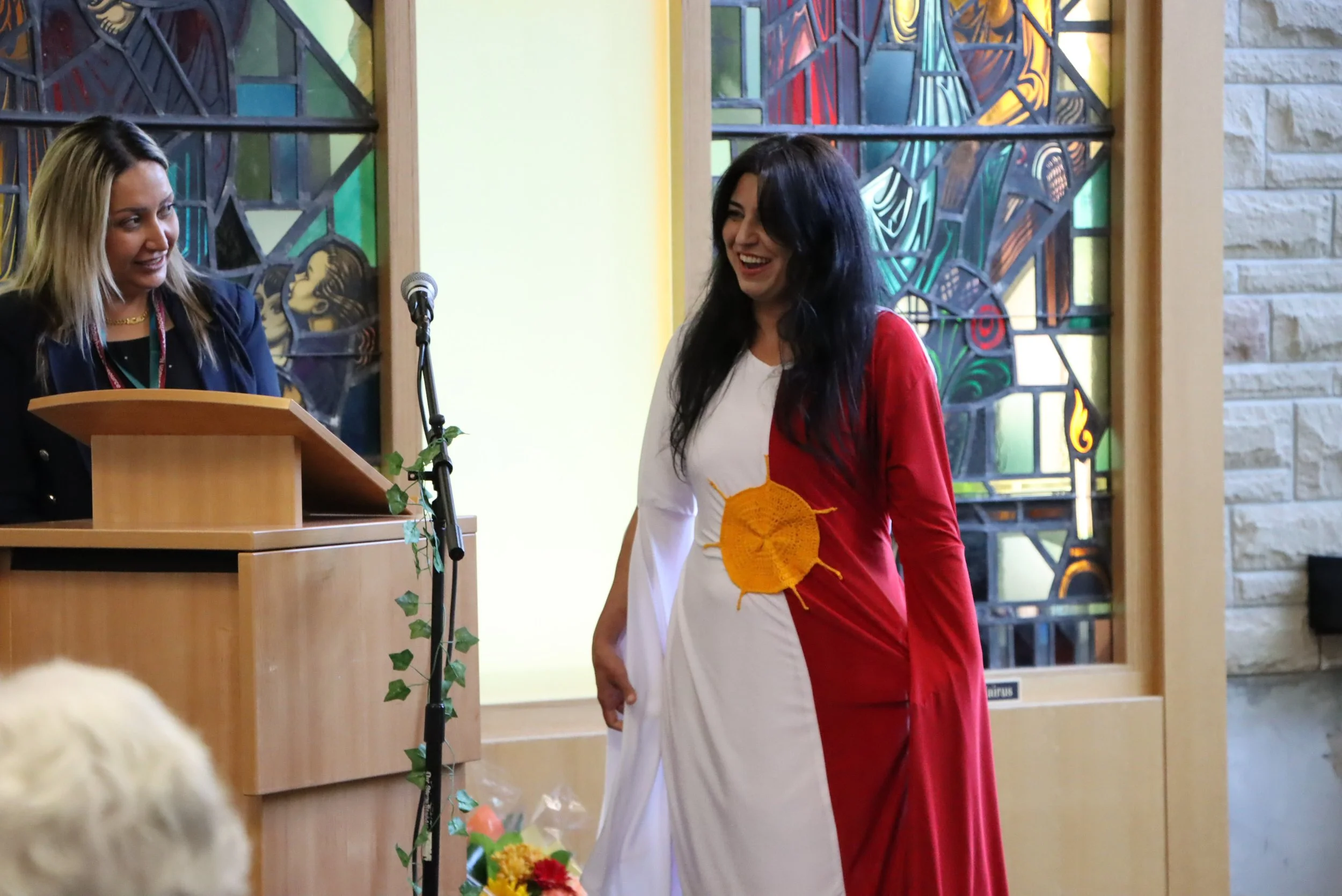 A woman dressed in a white and red dress symbolizing the Yazidi flag is smiling as she stands near a microphone. Another woman is at a podium, smiling and looking at her, with stained glass windows and stone walls in the background.