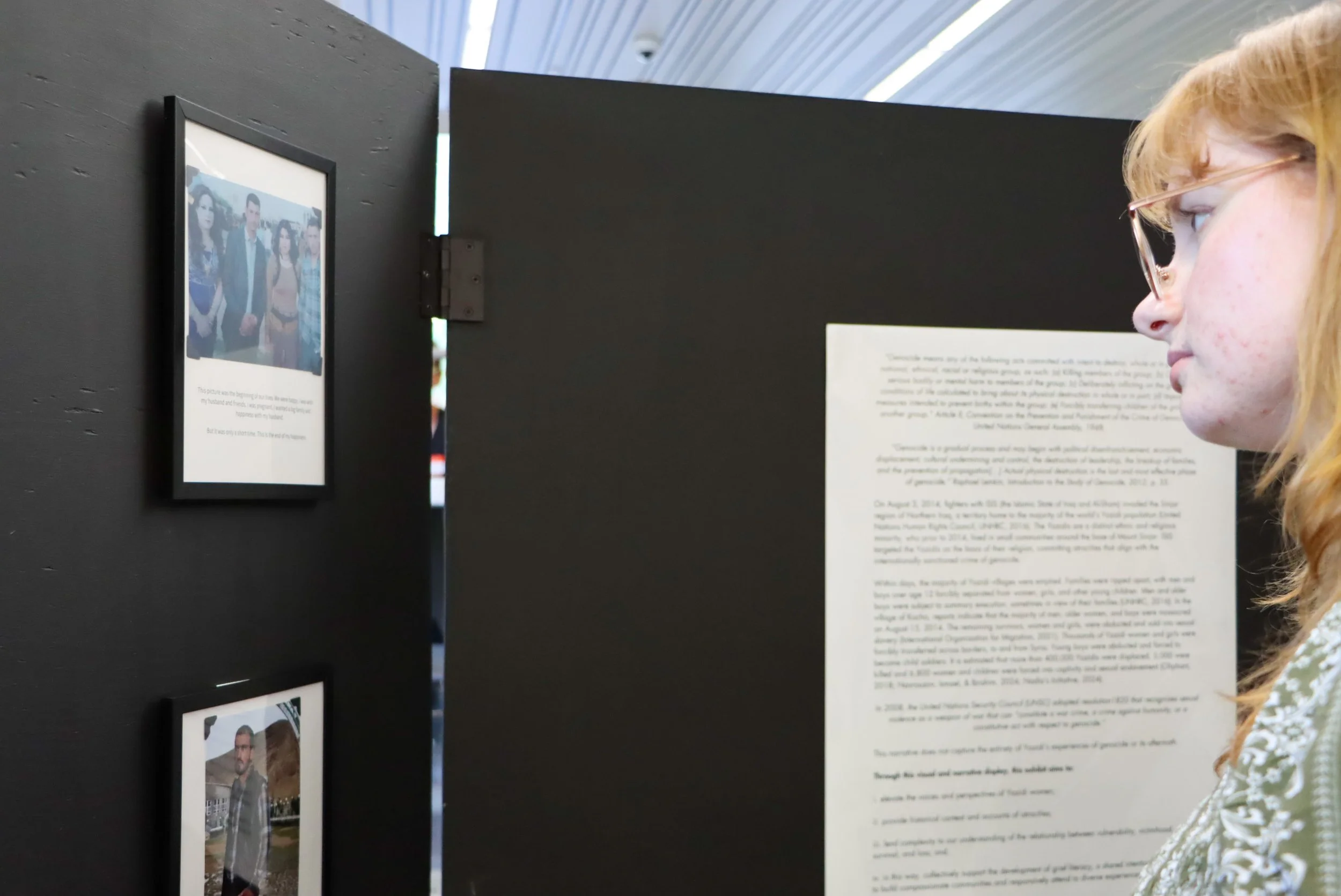 A woman with red hair and glasses viewing an exhibit with black display panels that hold photographs and text for the 2024 Yazidi Photovoice Exhibit held at King's University College. 