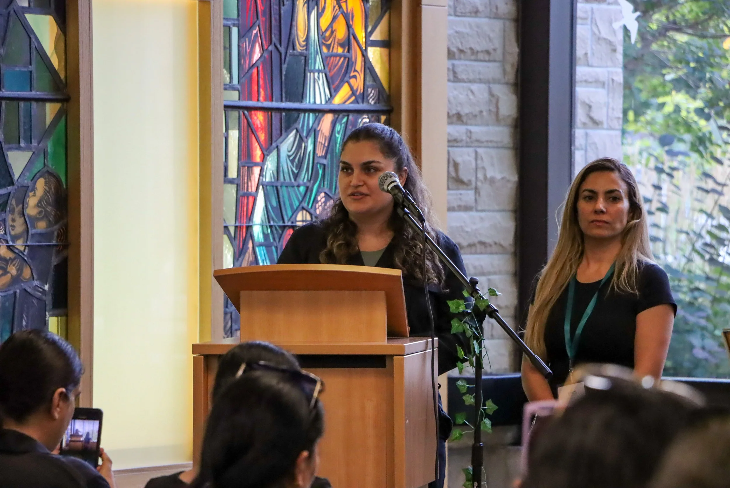 A woman with dark, curly hair, speaking at a podium with a microphone during a gathering, with another woman with blond hair standing beside her. The background features stained glass and stone walls, with windows showing green foliage outside. Audie