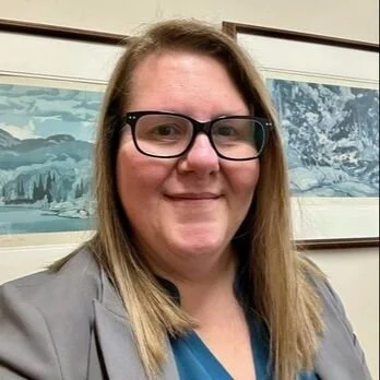 A woman with light brown hair wearing glasses, a gray blazer, and a blue top, smiling in front of framed artwork depicting snow-covered mountains.