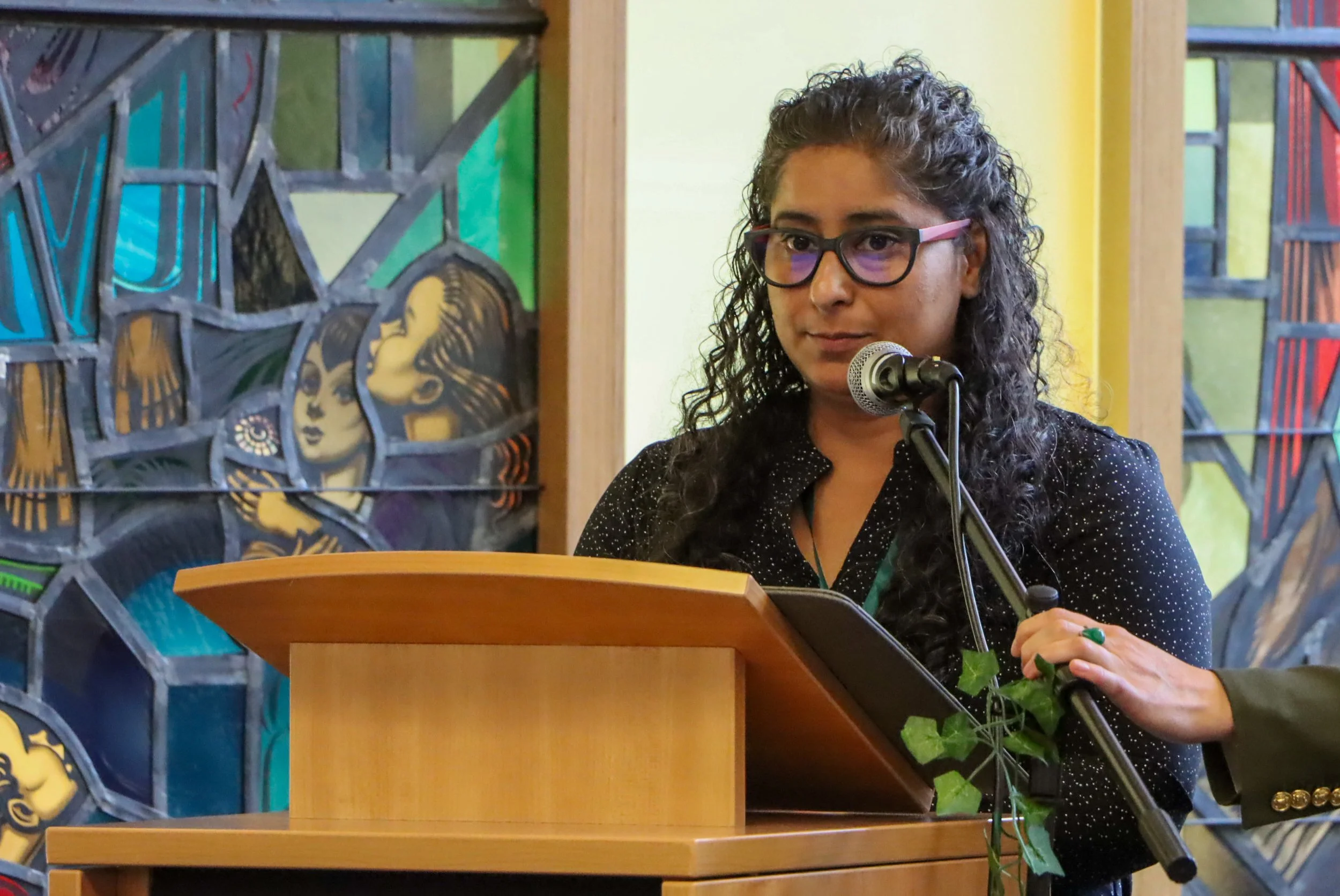 A woman with curly hair and glasses speaking at a podium during an event, with colorful stained-glass style artwork in the background.