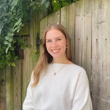 A young woman with long blonde hair smiling outdoors in front of a wooden fence and green foliage, wearing a white sweater and a necklace.