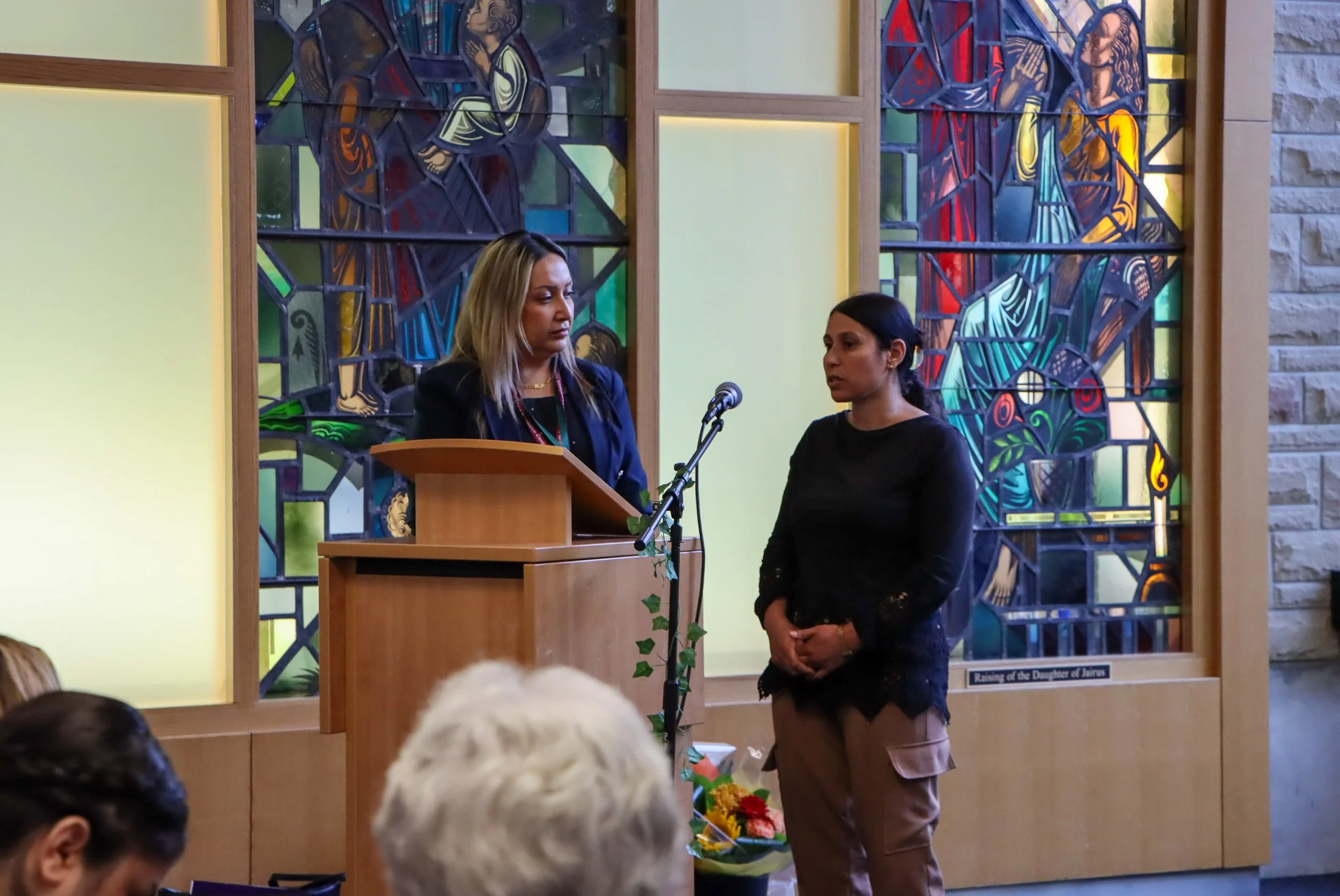 Two women standing near a microphone in front of stained glass windows in a church or religious setting. One woman is speaking at a podium, and the other is listening, with an audience visible in the foreground.