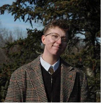 Person with short hair and glasses wearing a plaid blazer outdoors.