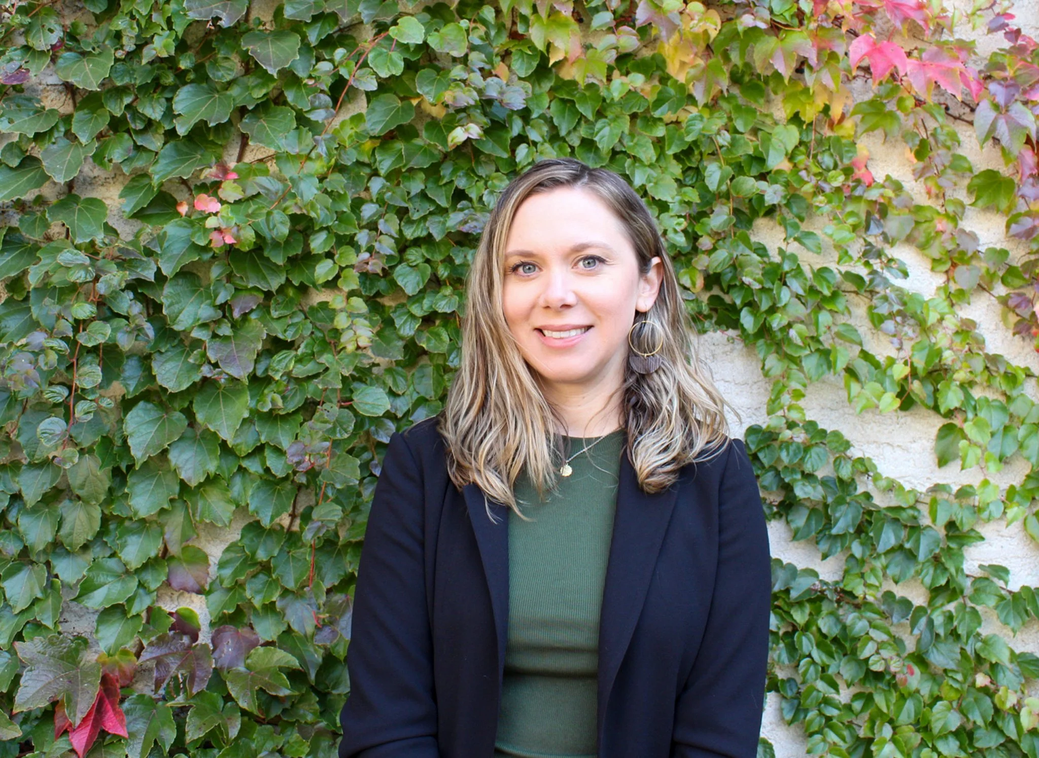 A woman with long, wavy blonde hair wearing hoop earrings, a green top, and a dark blazer, standing in front of a wall covered with green and pink ivy leaves.