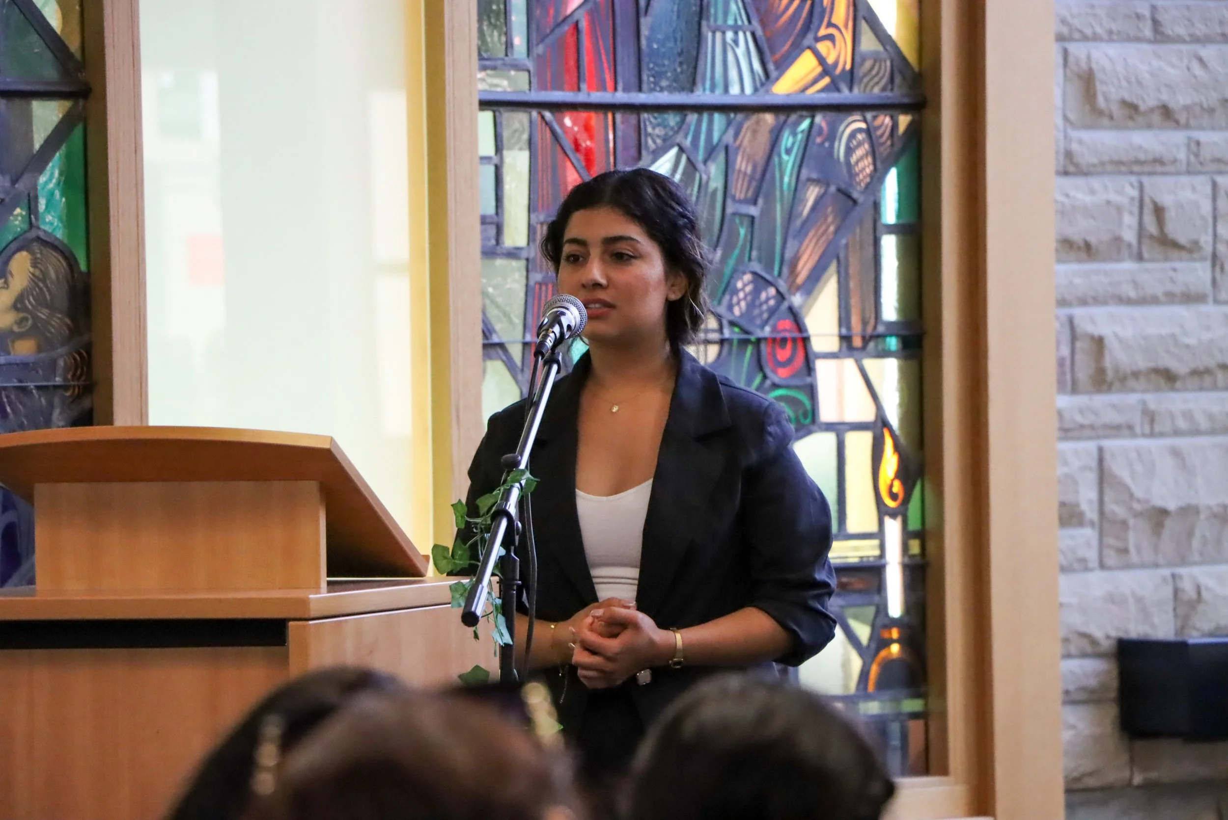 A woman with short dark hair, wearing a black blazer and white shirt, stands at a podium speaking into a microphone during the 2024 Yazidi Photovoice Exhibit at King's with colorful stained glass windows in the background.