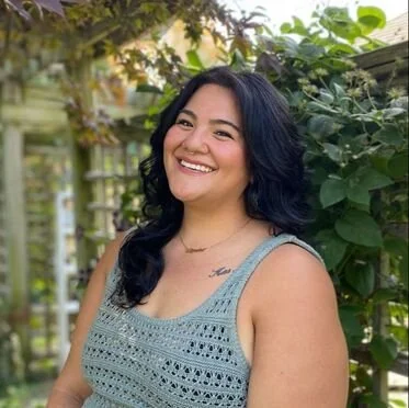 A woman with dark, wavy hair smiling outdoors with greenery and plants in the background.