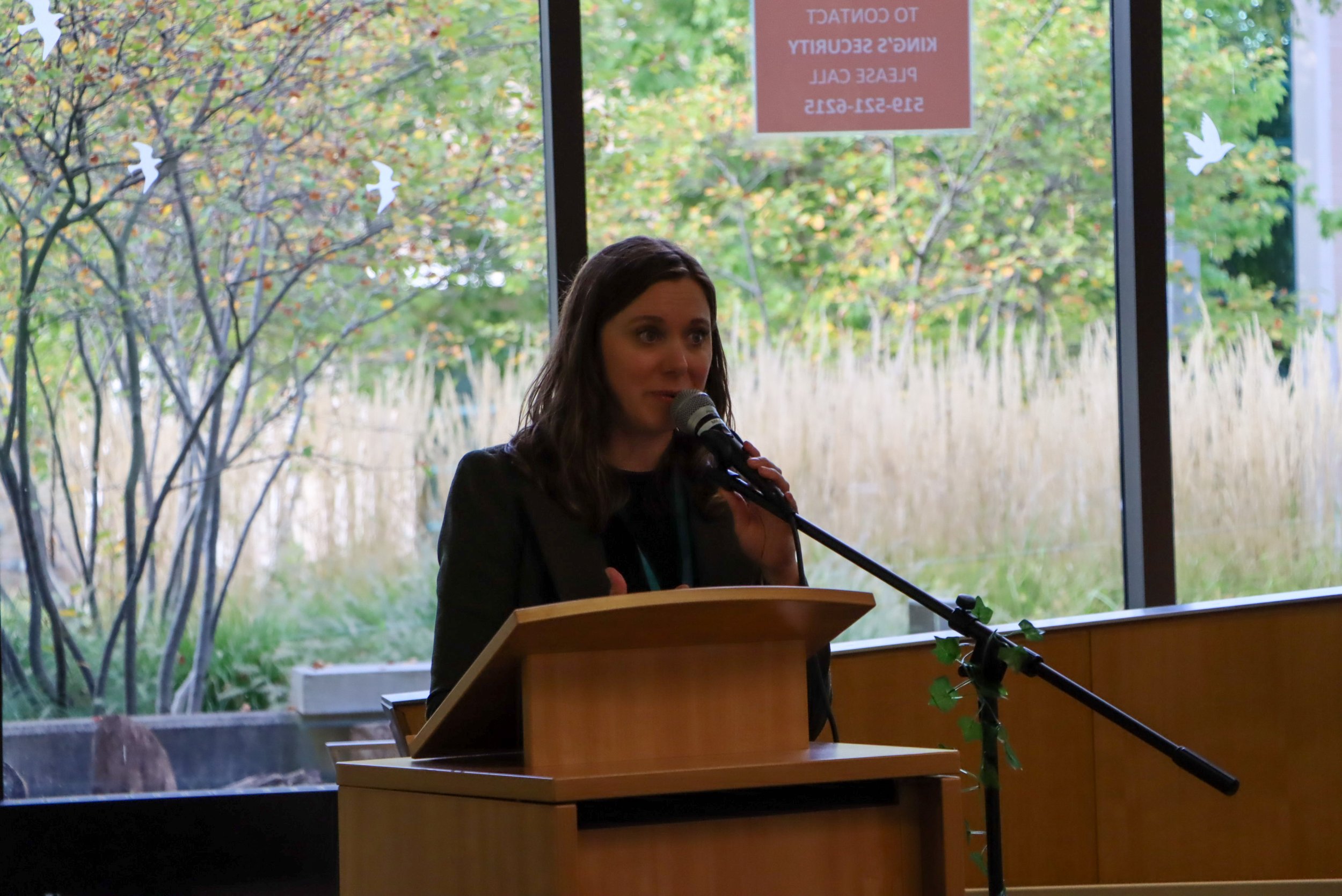 Woman speaking into a microphone at a podium with a backdrop of large windows showing trees with autumn foliage.