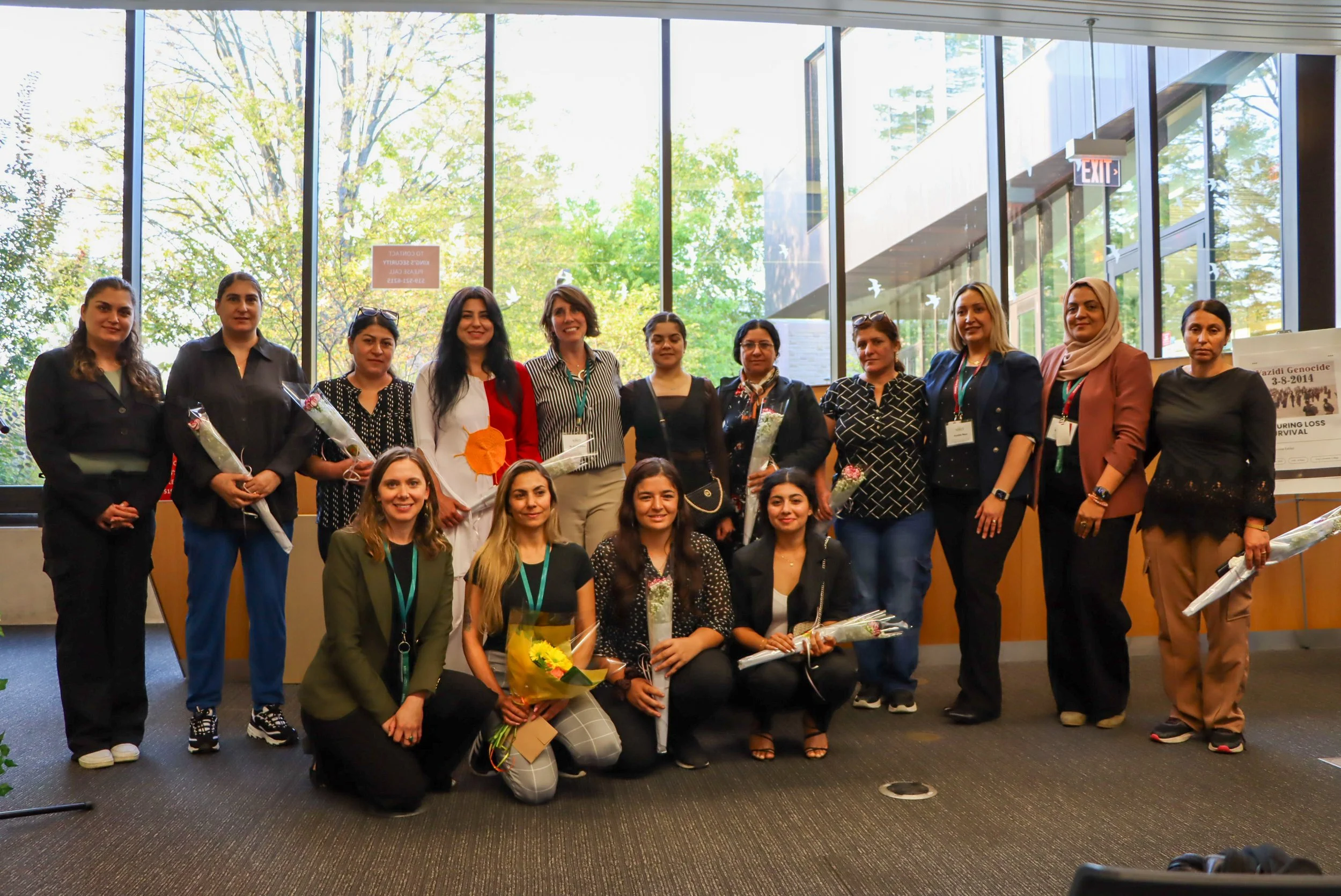 Group of women posing for a photo indoors near large window, some holding flowers and wearing conference badges.