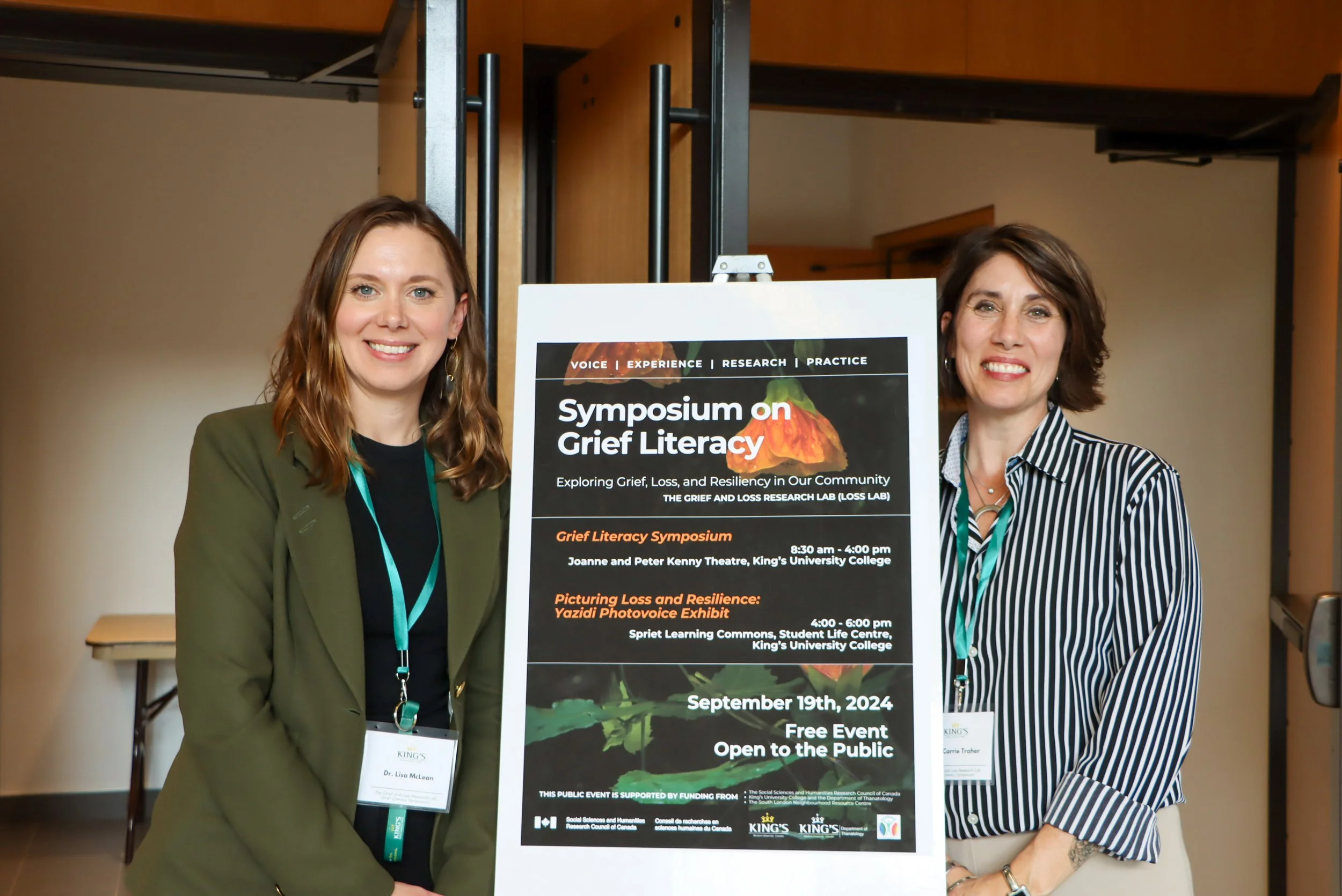 Two women standing next to each other smiling at an event. They stand near a poster that promotes a symposium on grief literacy with details about its schedule and location.