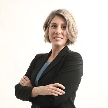 A woman with short, wavy gray hair wearing a black blazer and a blue top, standing against a white background.