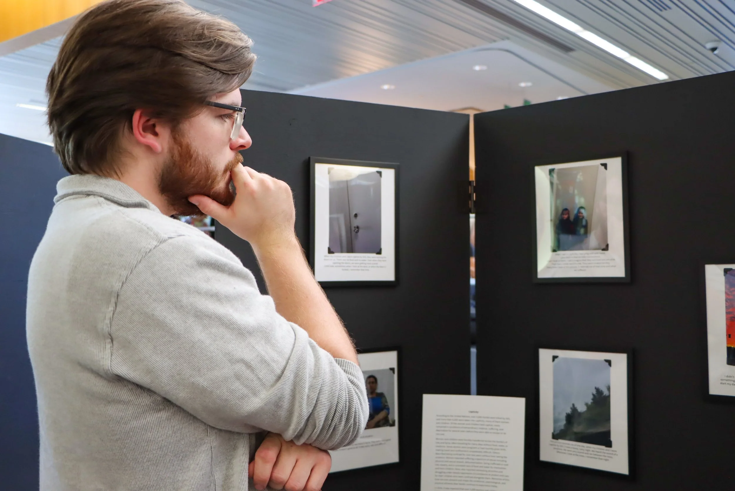 A man with glasses and a beard, wearing a light-colored sweatshirt, standing in front of framed photographs and text panels at the 2024 Yazidi Photovoice Exhibit held at King's University College. 