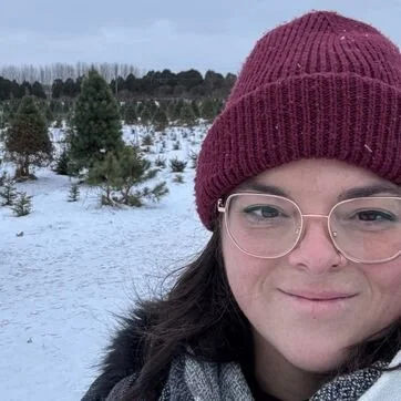Woman wearing a maroon knit hat and glasses in a snowy outdoor landscape with trees.