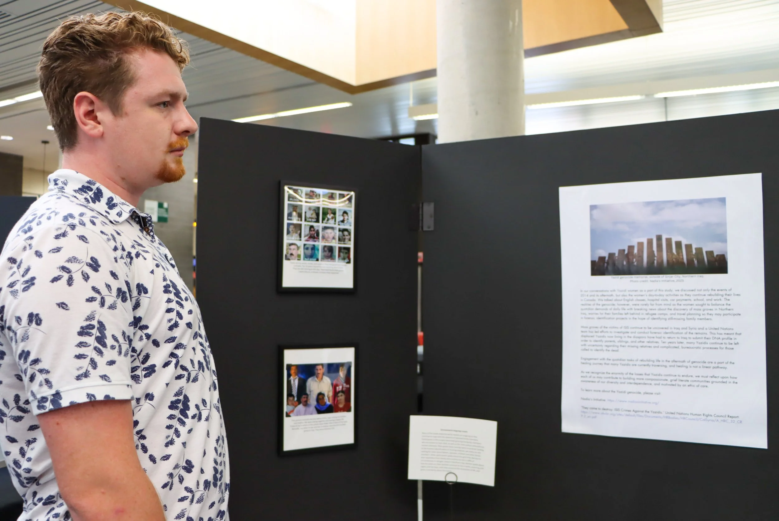 A young man with curly red hair and a beard, wearing a white shirt with a blue palm tree pattern, is looking at exhibition displays for the 2024 Yazidi Photovoice Exhibit held at King's University College. 