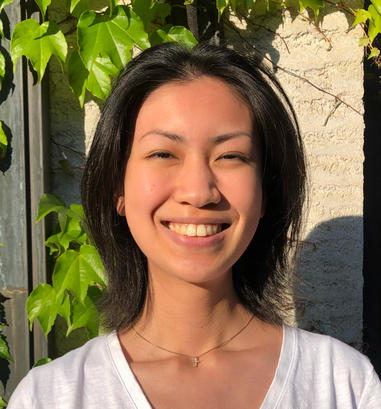 A young woman with black hair smiling outdoors in sunlight, with green leaves and a stone wall in the background.