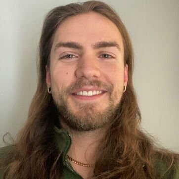 A young man with long brown hair, a beard, and earrings, smiling at the camera against a plain background.