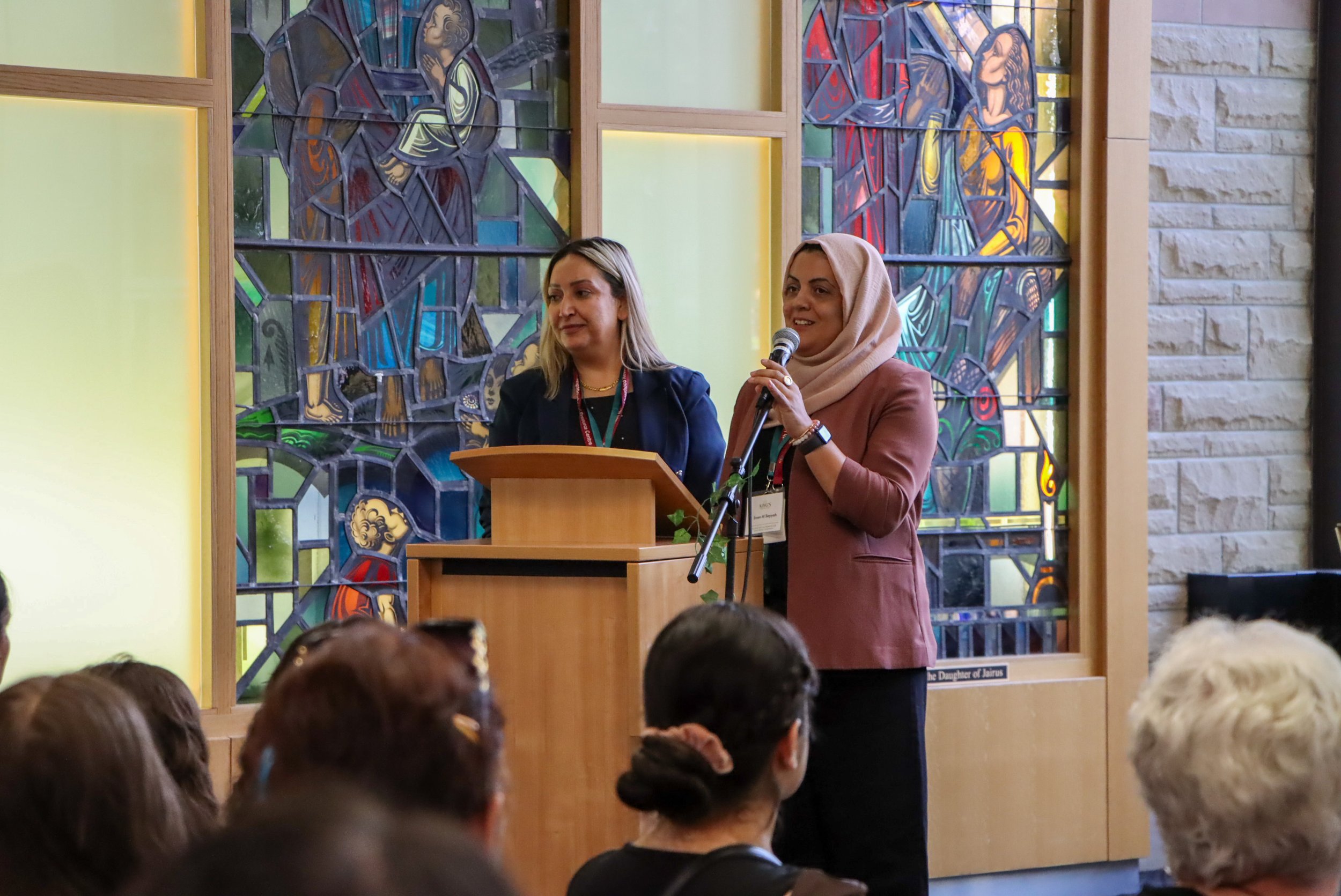 Two women are speaking at a podium in front of a stained glass window. The woman on the right is wearing a headscarf and brown blazer, holding a microphone. The woman on the left has blonde hair and is wearing a dark blazer. Audience members are visi