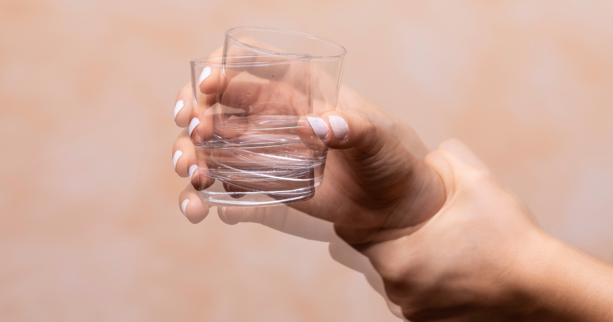 A hand holding a clear glass of water against a neutral background.