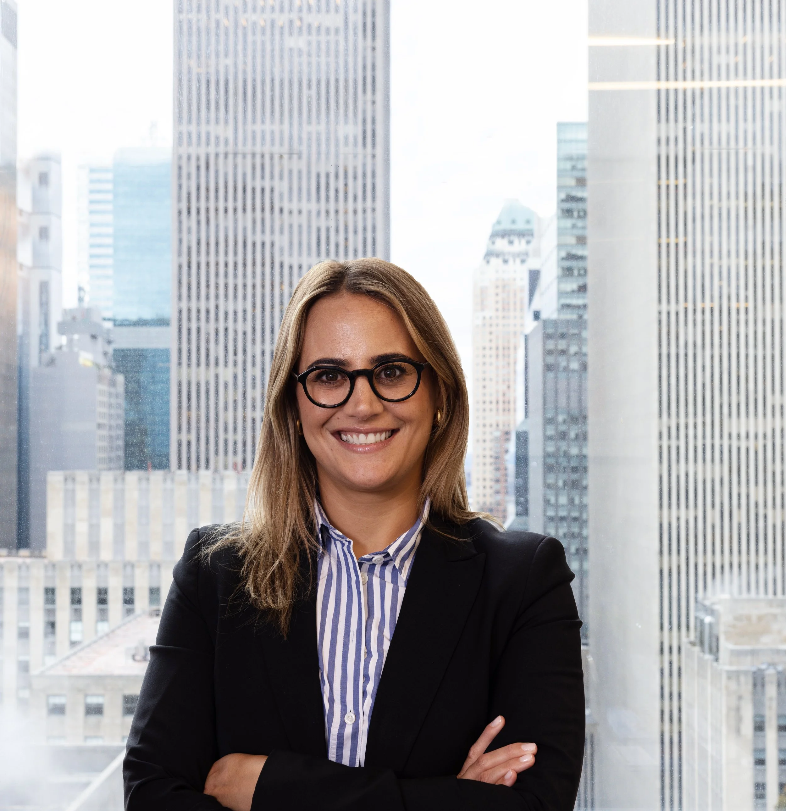 Professional woman with blonde hair and glasses, smiling, standing with arms crossed in front of large office windows showing city skyscrapers.