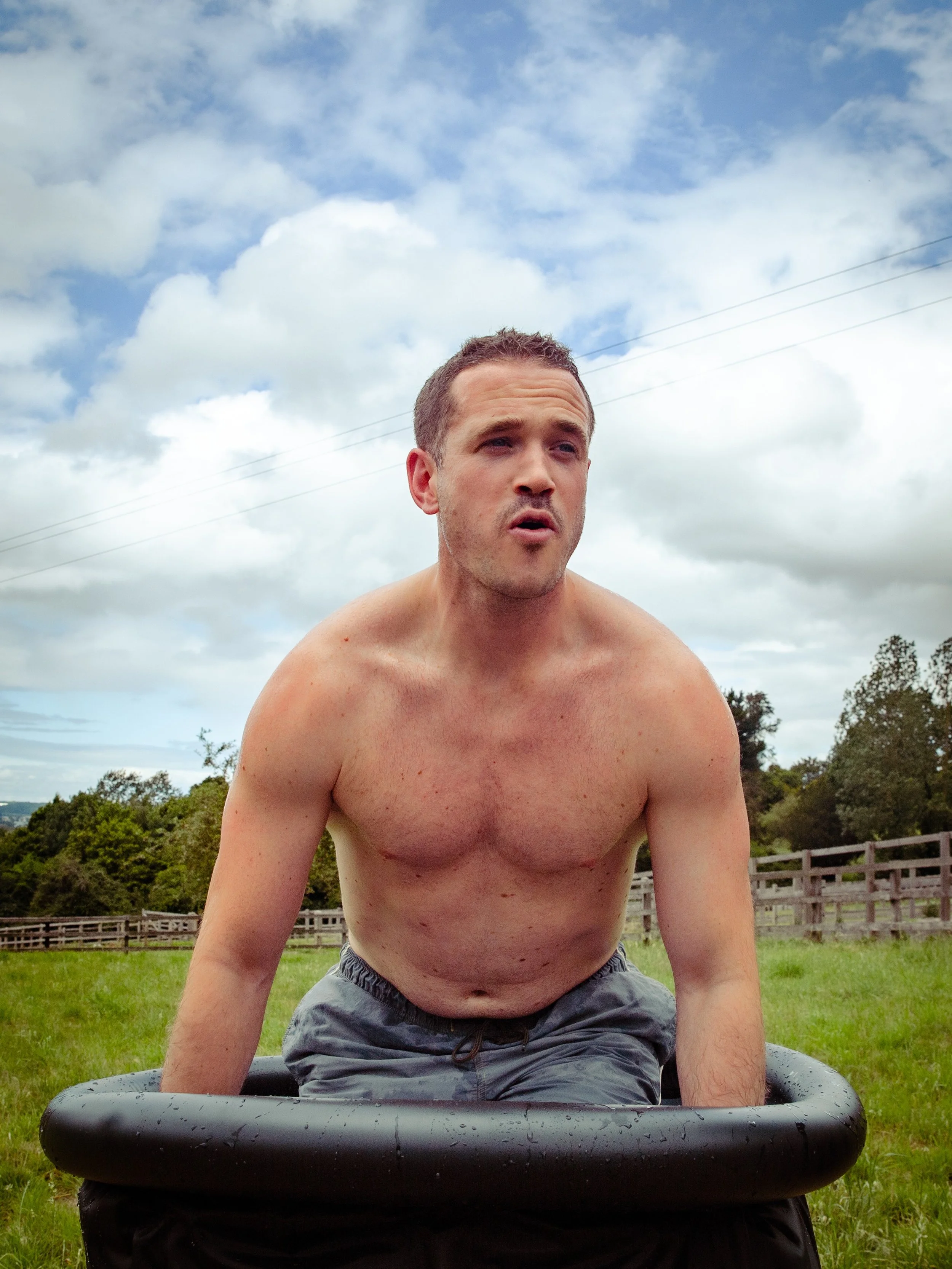 A shirtless man with short hair kneeling outdoors on a sunny day, with a cloudy sky, trees, and a wooden fence in the background.