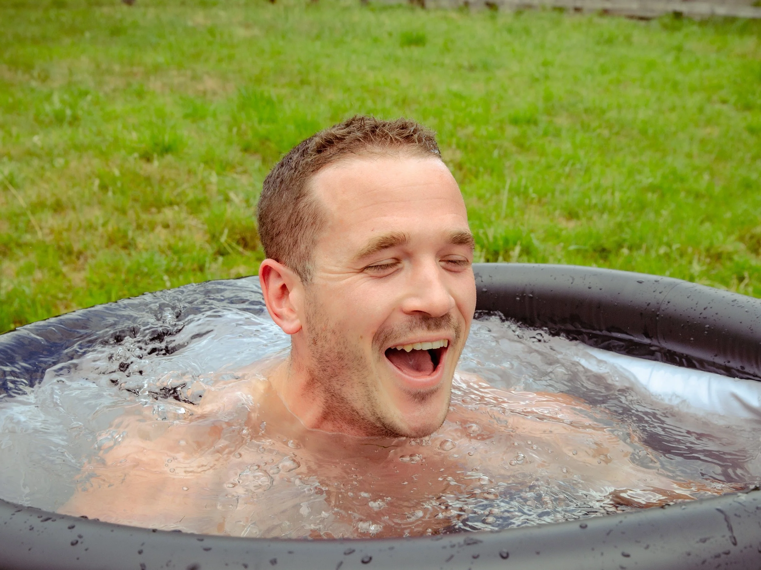 A man smiling and enjoying a soak in an outdoor hot tub with lush green grass in the background.