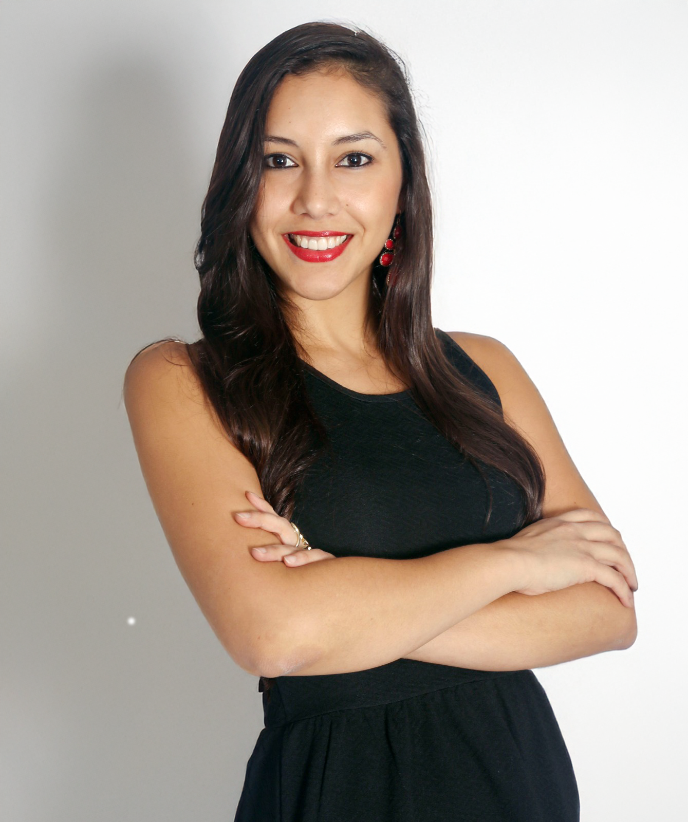 A young woman with long dark hair, red lipstick, wearing a sleeveless black dress, smiling with arms crossed, standing against a plain background.