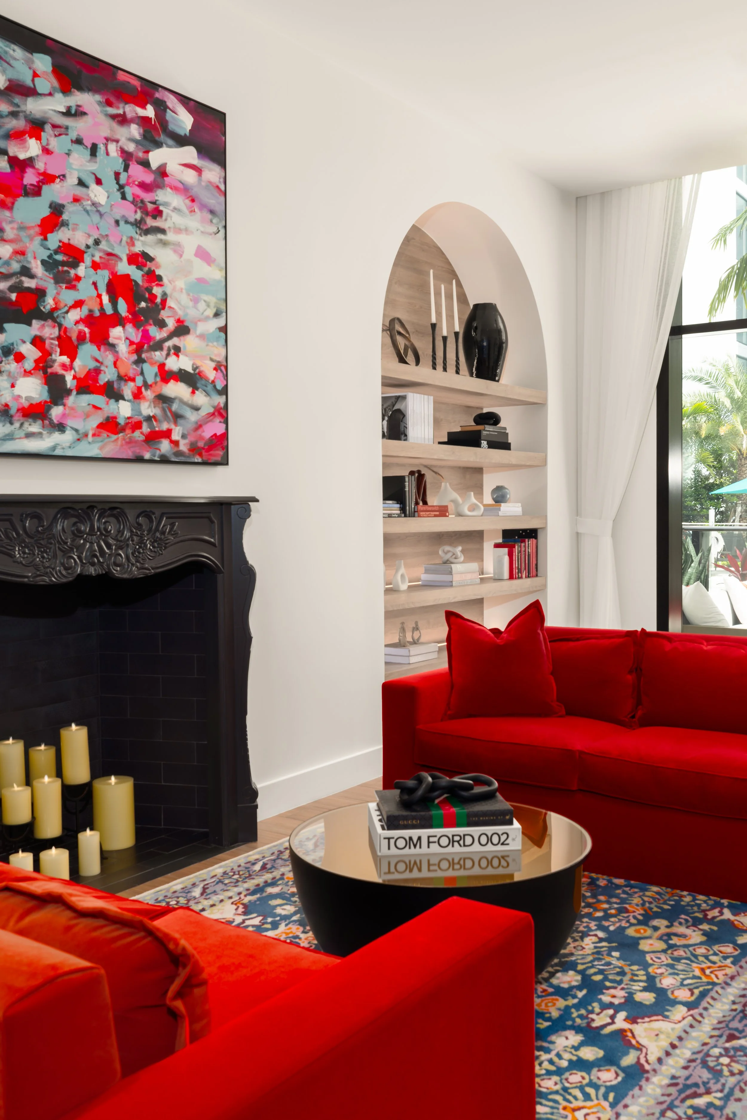 A cozy living room with a red velvet sofa, a black fireplace with candles inside, a colorful patterned rug, a round coffee table with fashion books including Tom Ford, and a modern white bookshelf with decorative objects, a window with white curtains, and a leafy plant outside.