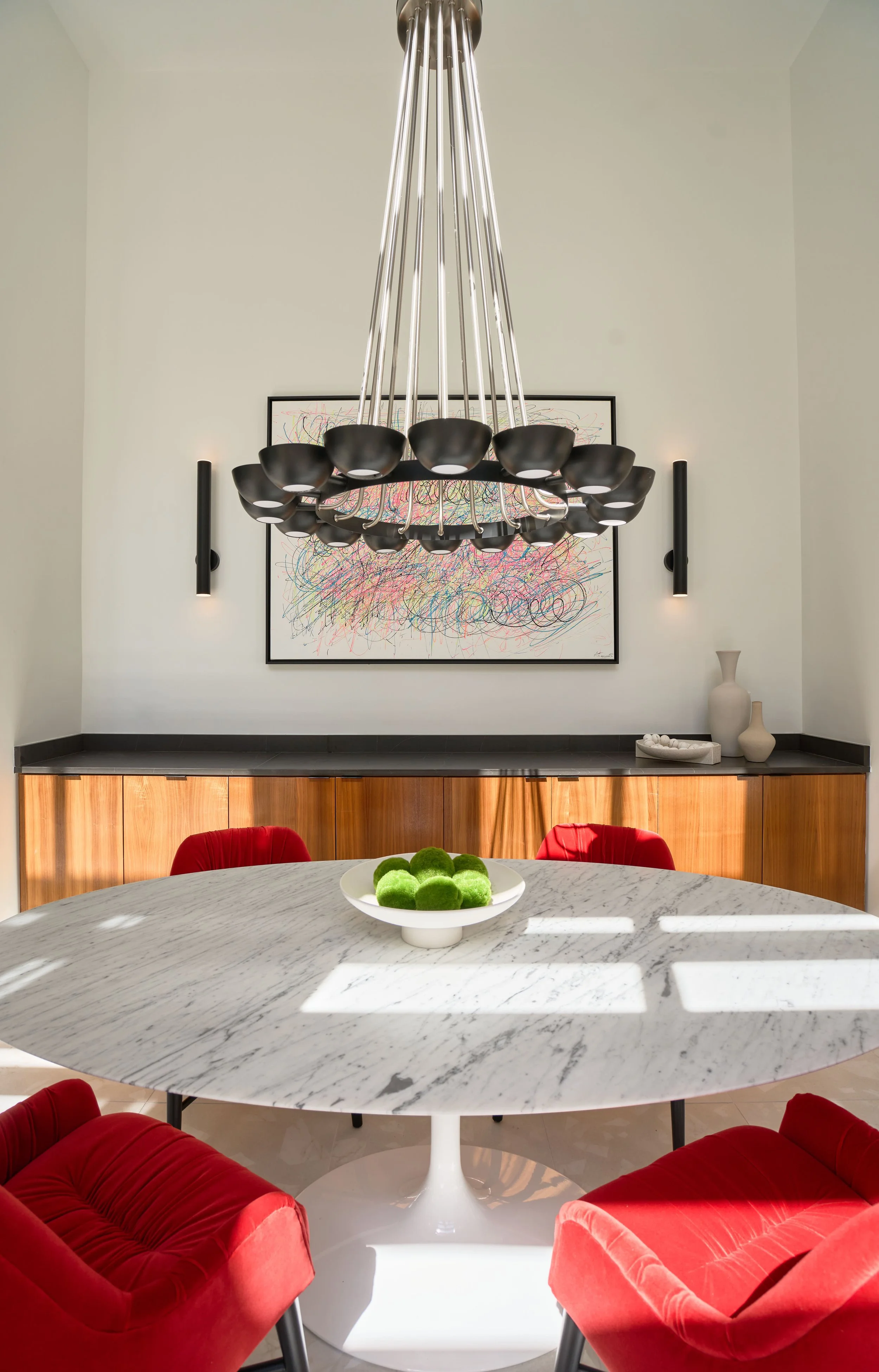 A dining room with a marble round table, surrounded by red velvet chairs, a modern chandelier hanging above, and a black and white abstract painting on the wall. There are vases and a plate of green decorative objects on the sideboard.
