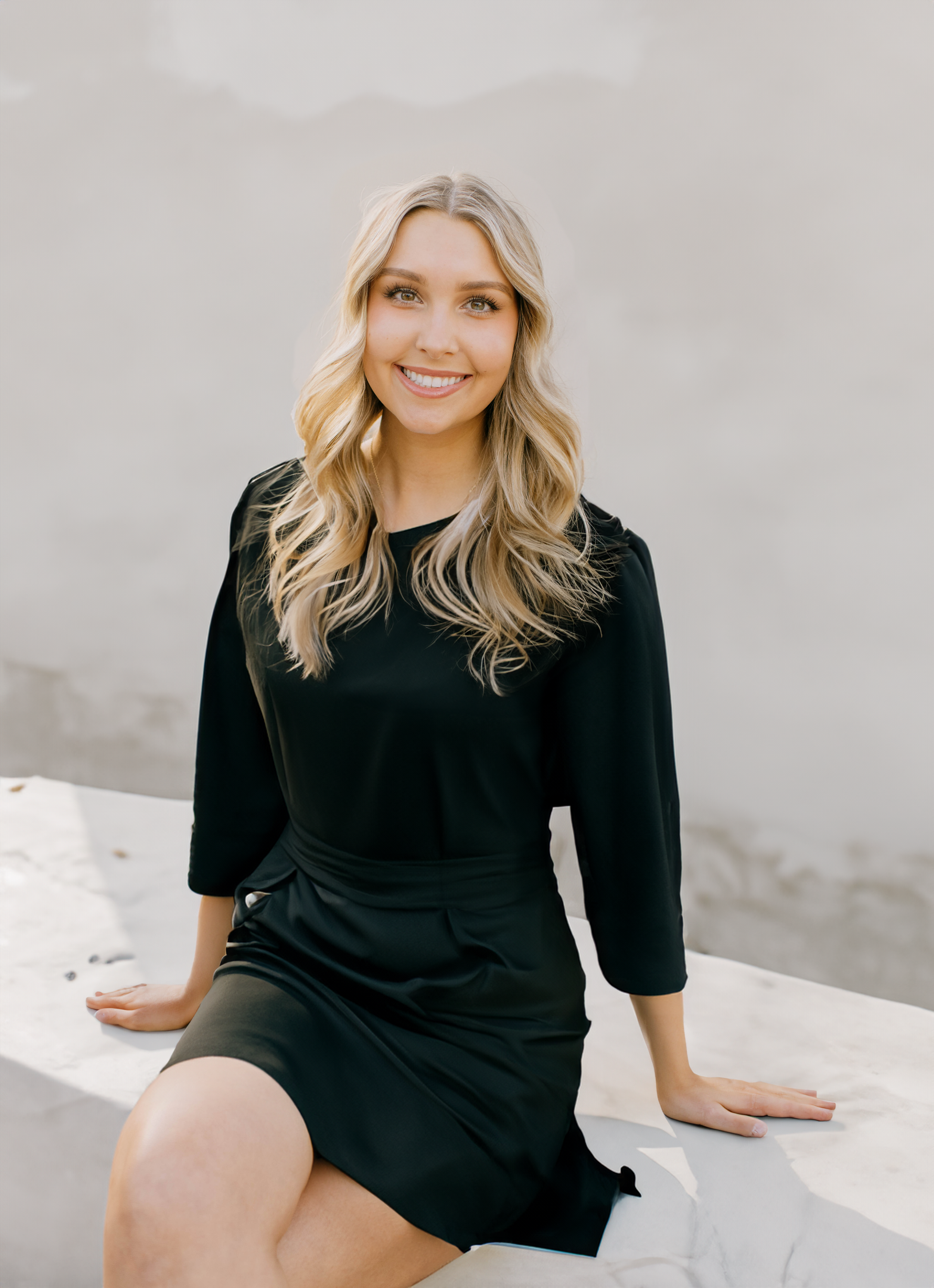 A young woman with blonde wavy hair, smiling, sitting on a white surface at the beach, wearing a black dress.