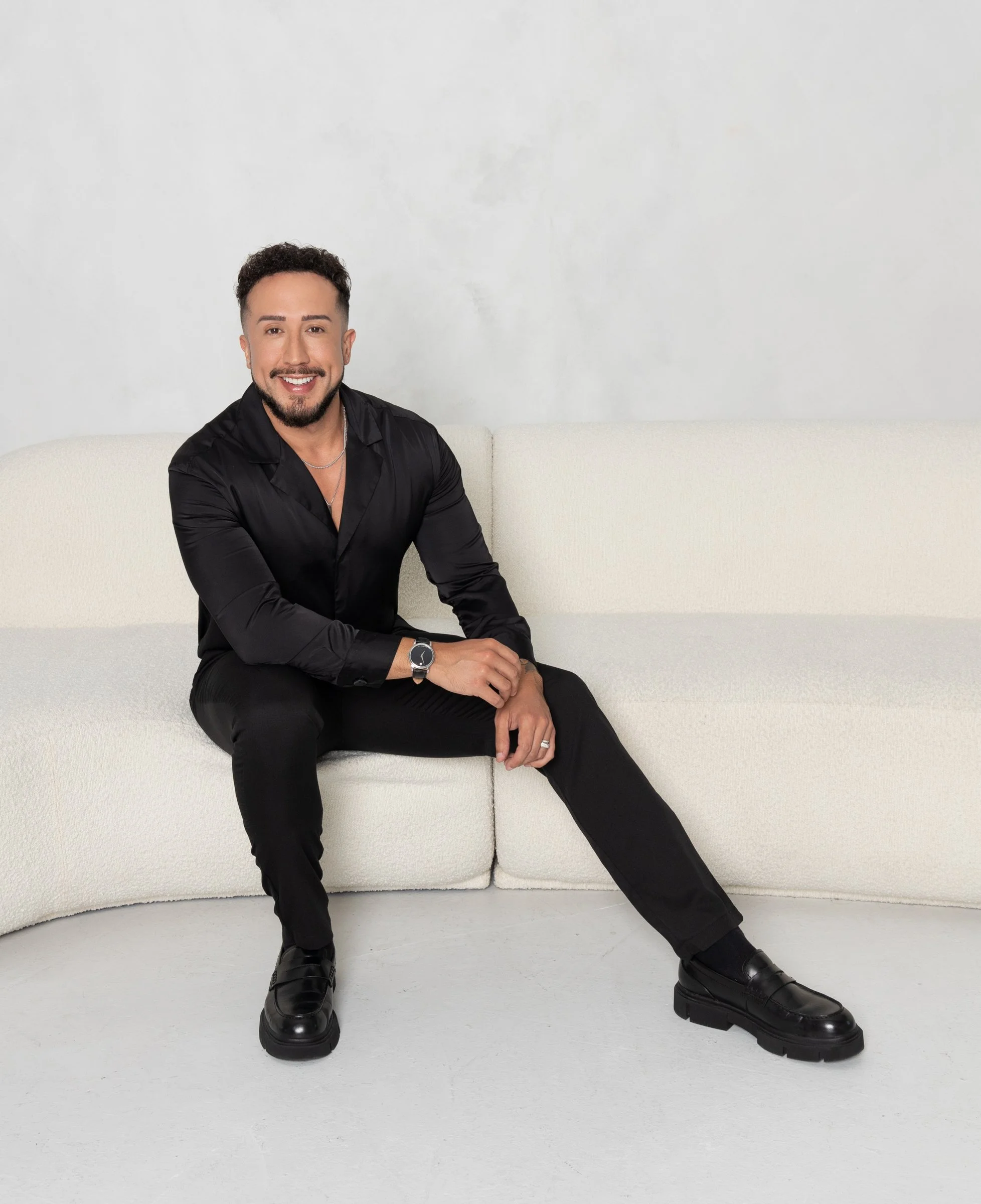 A man with short, dark curly hair, a beard, and mustache, wearing a black shirt, black pants, and black shoes, is sitting on a white sofa against a plain white wall. He is smiling and looking at the camera with his left arm resting on his lap.