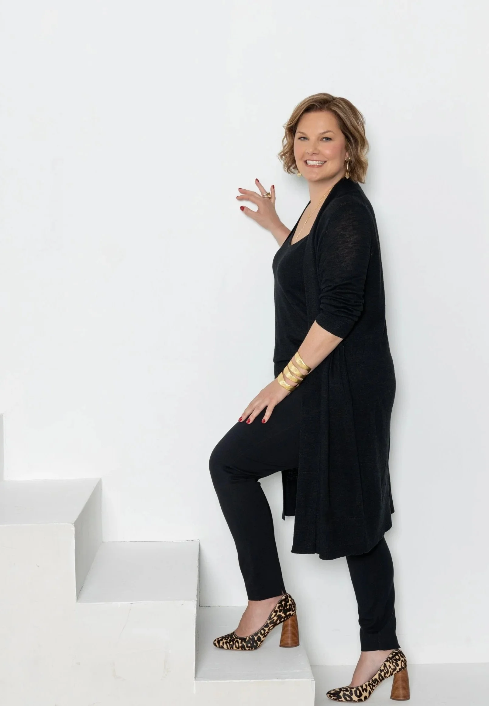 A woman with short wavy hair is smiling and posing against a white wall. She is wearing a black long-sleeve dress with a slit, gold jewelry, and leopard-print high heels. She is standing on stair steps with her left foot on a higher step, touching the wall with her right hand.