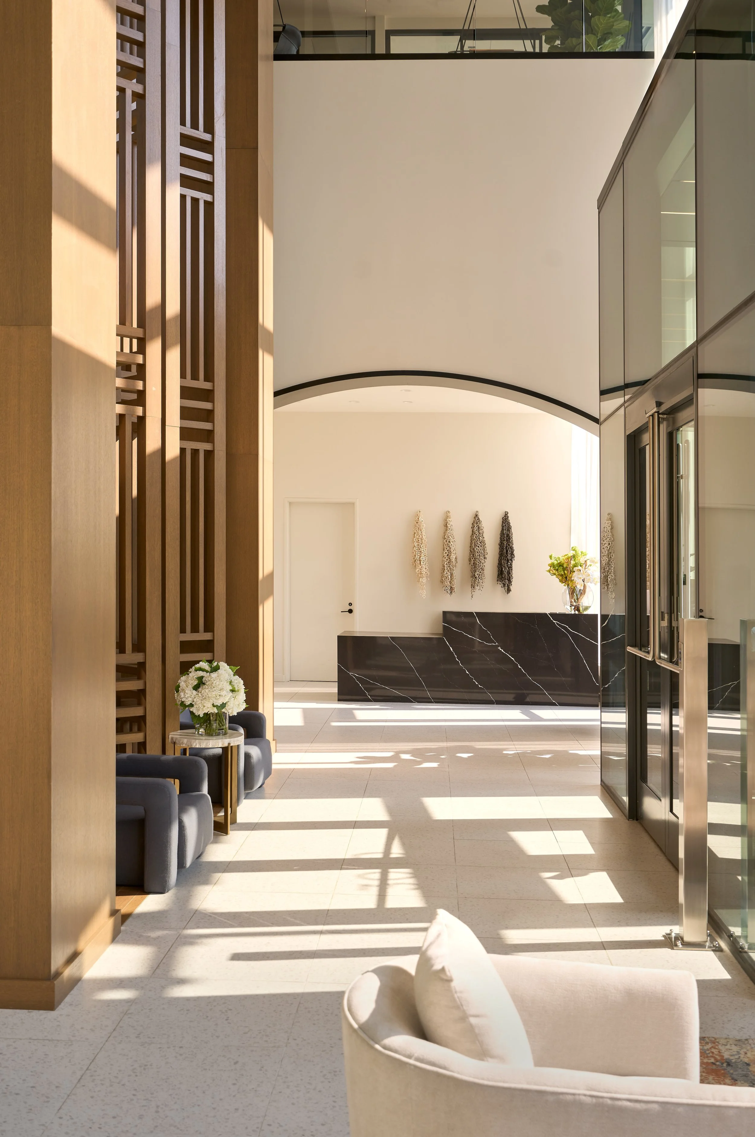 Modern hotel interior with white walls, beige floor tiles, and large windows letting in natural light. There is a white couch with a pillow, a small side table with white flowers, and a black marble reception desk with decorative wall art.