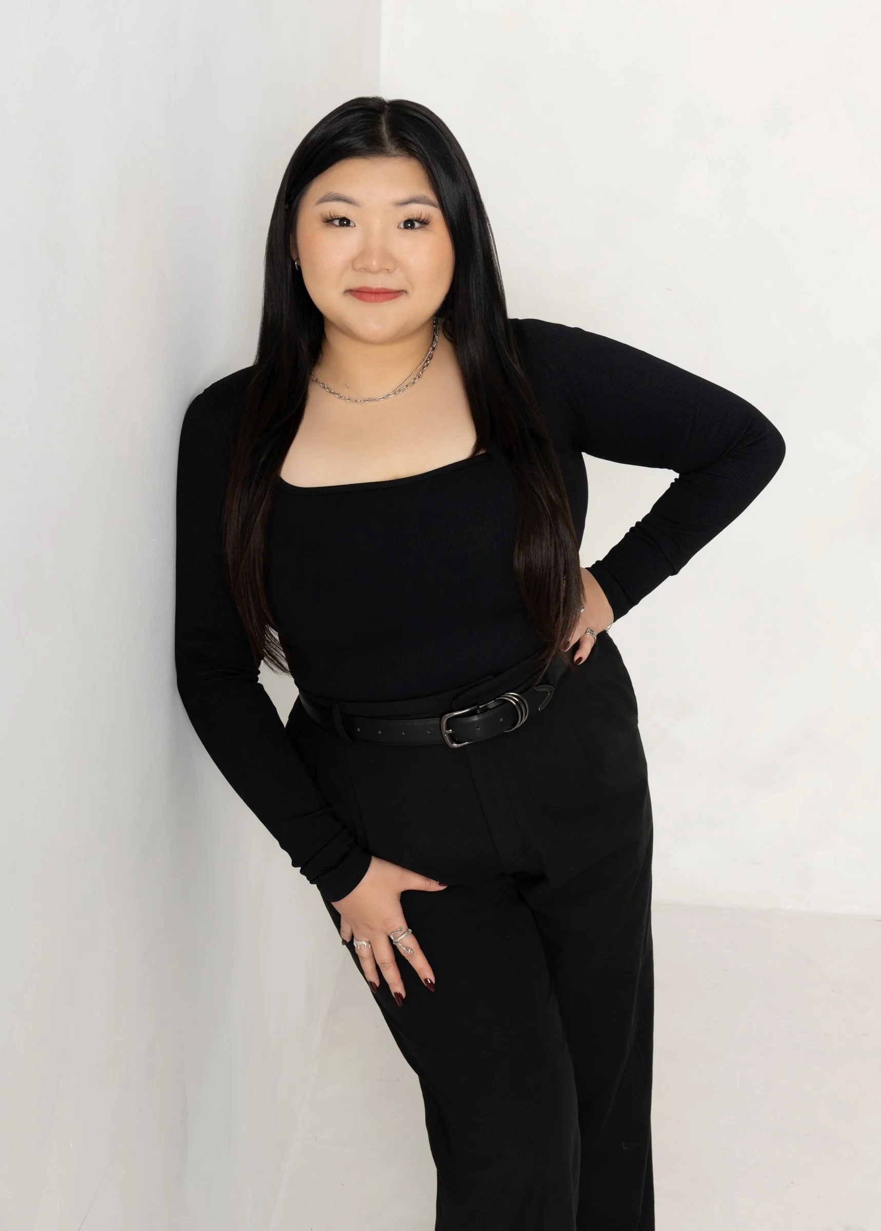 A woman with long black hair wearing a black long-sleeve top and black pants, standing against a white wall, posing with her right hand on her hip and her left hand resting near her thigh.