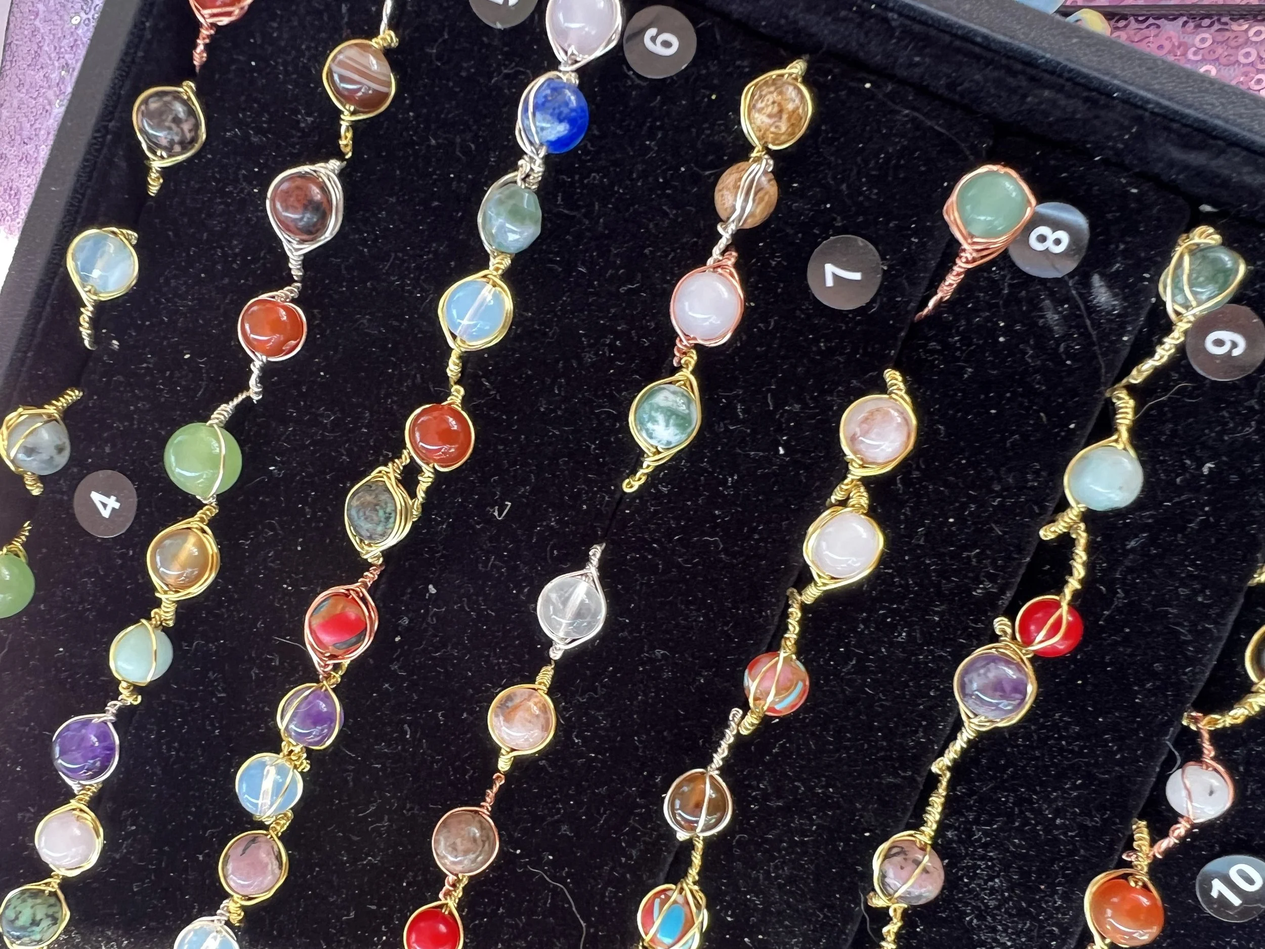 Colorful gemstone rings with gold, silver, and copper bands displayed on a black velvet tray, each ring labeled with a small black and white numbered tag.