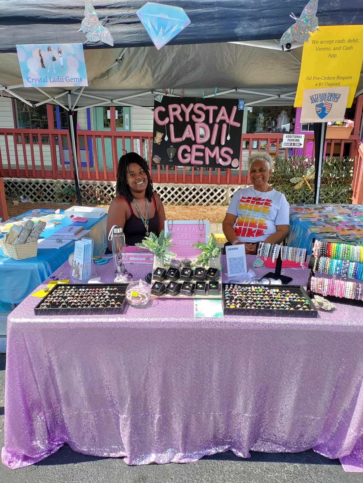 A jewelry booth at an outdoor market with two women sitting behind the table. The table is covered with a purple, glittery tablecloth displaying rings, bracelets, and jewelry pieces. Signs include 