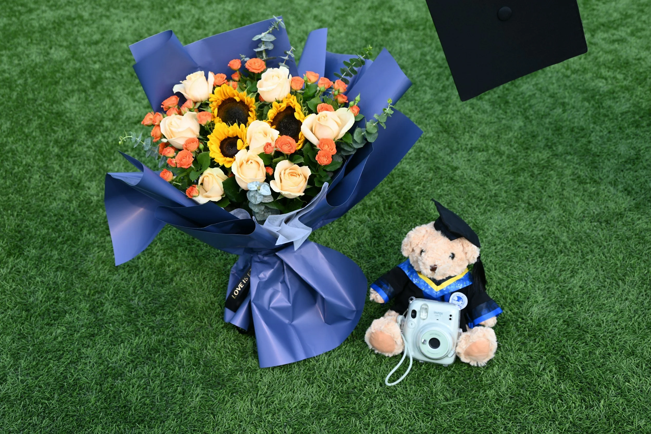 A bouquet of flowers with white roses, sunflowers, and orange small roses wrapped in blue paper, placed on green grass. Nearby, a teddy bear wearing a graduation gown and cap, holding a camera, is sitting on the grass.