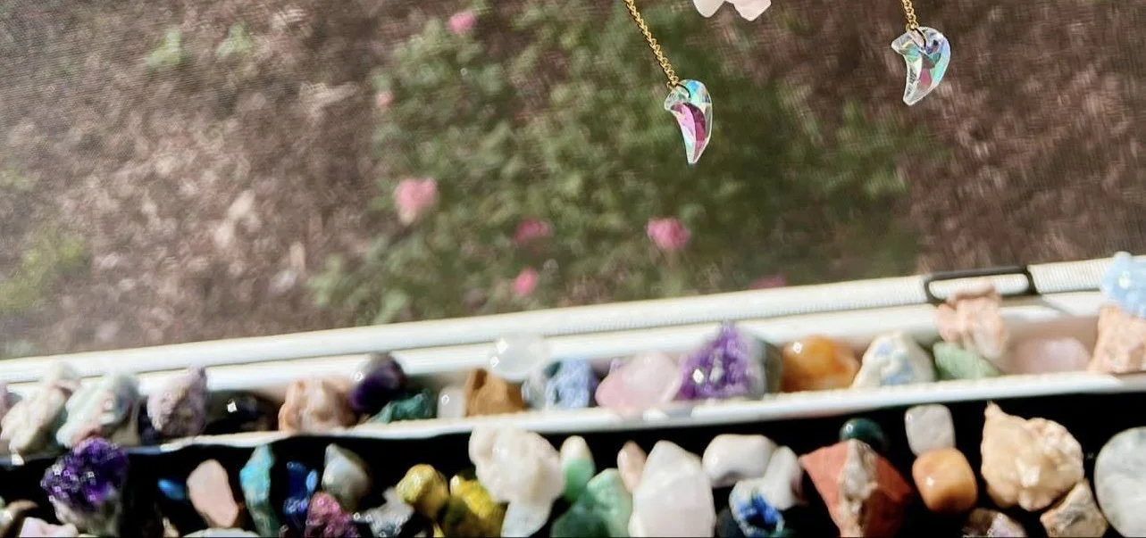 Close-up of a windowsill with various colorful rocks and crystals, with a blurred view of a garden with pink flowers outside.