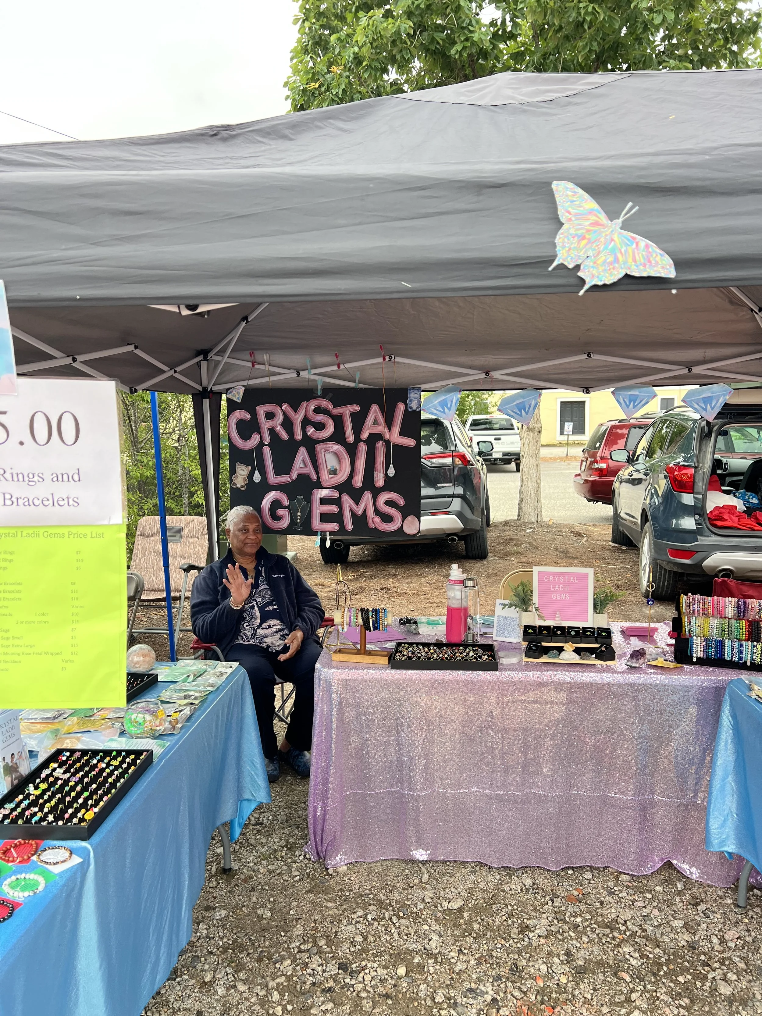 A woman sitting at a booth selling jewelry and crystals at an outdoor market. The booth is decorated with pink and blue tablecloths, with a sign reading 'Crystal Ladii Gems' and various jewelry displays. There are parked cars and trees in the backgro