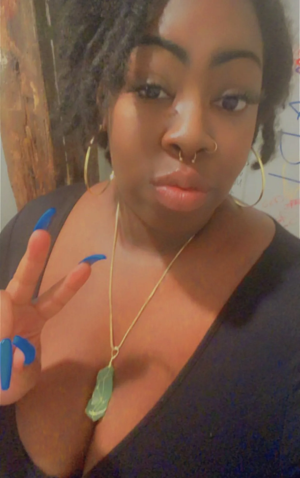 A woman with dark hair, wearing gold hoop earrings and a gold necklace with a pendant, making a peace sign with her blue painted nails. She has a nose ring and is partially undressed, with a black top draped over her shoulders, against a background w