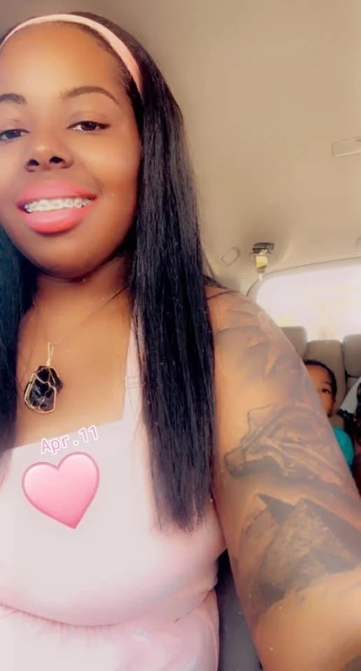 A woman taking a selfie on a bus, smiling with braces, wearing a pink headband and a necklace with a black stone. She has long black hair and a large tattoo on her arm. A child is partially visible in the background.