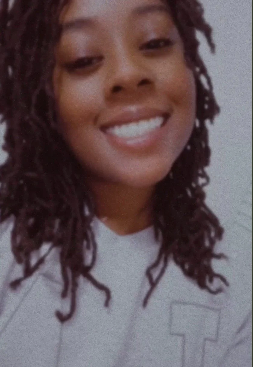 Close-up photo of a woman with dark, curly hair smiling showing her teeth, wearing a light-colored shirt.