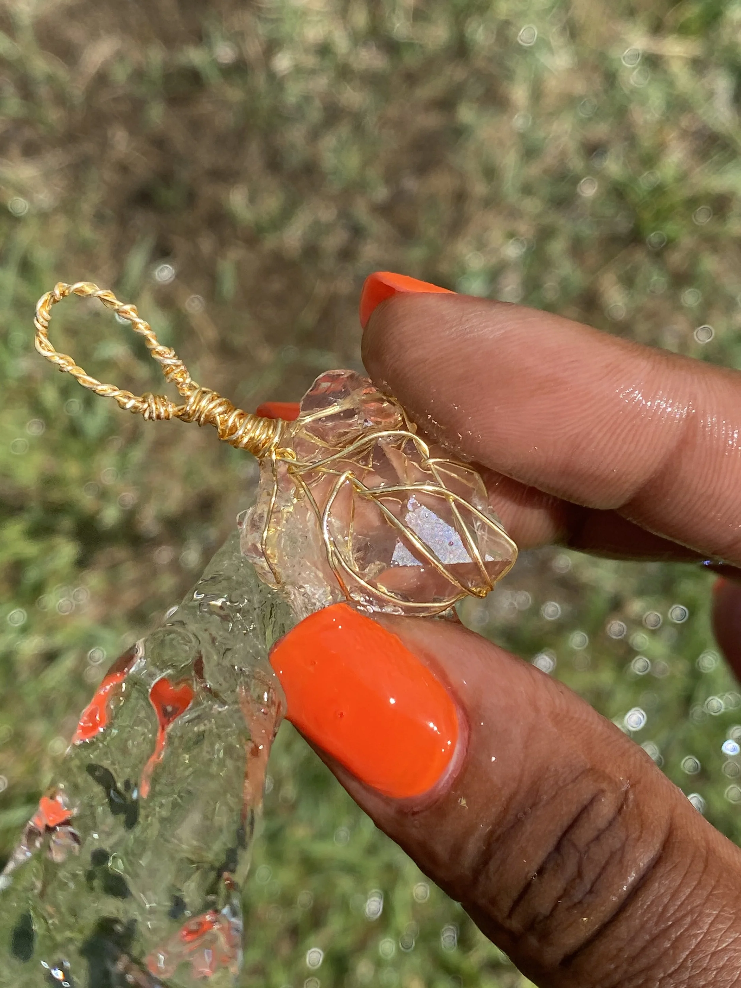 Close-up of a person holding a small, clear crystal with gold wire wrapping, outdoors on grass and water droplets visible.